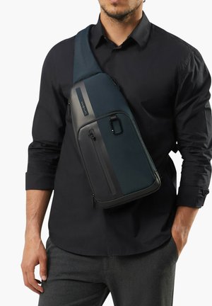 Man wearing black shirt and gray pants with navy and black crossbody sling bag featuring zippered compartments.