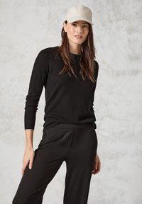 Svart långärmad topp med ett strukturerat mönster, kombinerad med svarta byxor. En beige keps fulländar looken, mot en neutral bakgrund.