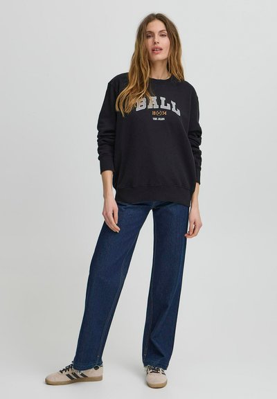 Fekete kapucnis pulóver "BALL 1974 THE JEANS" felirattal, széles lábú farmernadrággal párosítva. A modellen bézs sportcipő van, sötét csíkokkal.