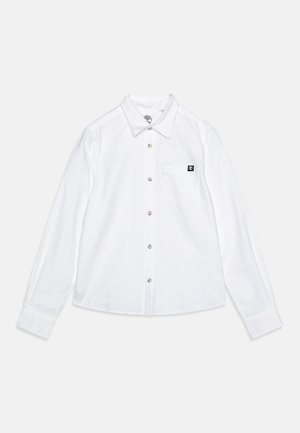 Chemise blanche à manches longues en coton. Elle possède un col pointu, une fermeture à boutons sur le devant, une poche poitrine et un subtil patch de marque. Texture lisse.