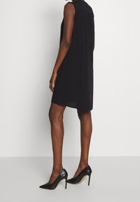 Kvinna i en ärmlös svart klänning och svarta högklackade skor som står mot en enkel vit bakgrund.