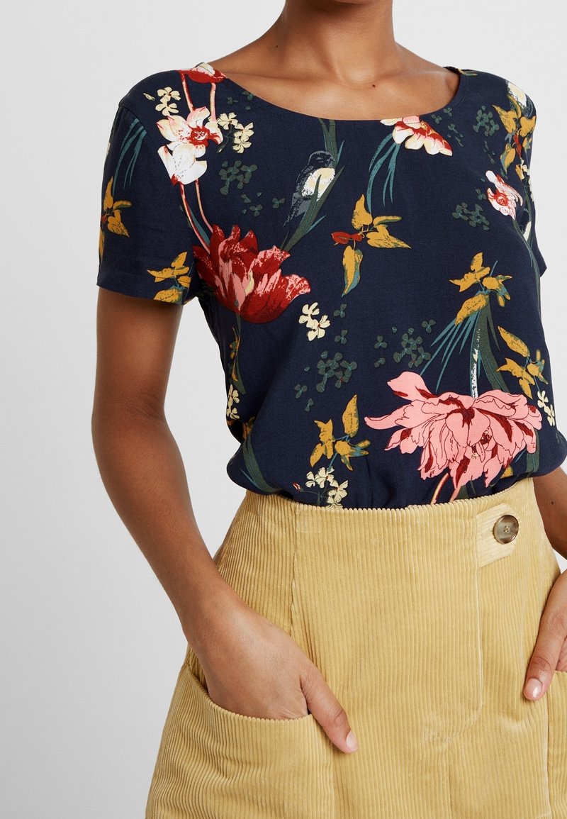 Granatowa bluzka z krótkim rękawem w kwiatowy wzór, z czerwonymi, różowymi i żółtymi kwiatami na ciemnym tle, zestawiona z teksturowaną spódnicą z beżowego sztruksu.