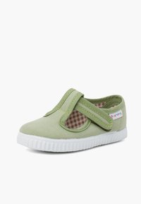 Zapatilla de lona verde con una suela de goma blanca texturizada, correa de Velcro y detalles recortados en los laterales que revelan un patrón de cuadros en el interior.