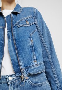Modrá denimová bunda s krátkým střihem, předním zipem, dvěma náprsními kapsami a jemným opotřebením. Nositelná přes bílou košili.