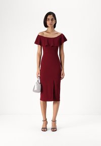 Burgundské off-shoulder šaty s volánovým výstrihom, priliehavým strihom, dĺžkou po kolená a bočným rozparkom. V páre so striebornou kabelkou a sandálmi na podpätku.