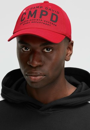 Red cap with black text "CMPD" and additional branding, paired with a black hooded sweatshirt. Simple design, structured shape, cotton material.