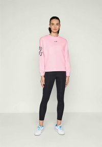 Felpa rosa con la scritta "goodness" stampata in nero sulla manica sinistra, abbinata a leggings neri. Sneakers bianche con accenti blu completano l'outfit.