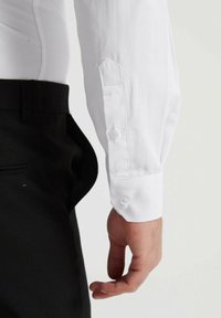 Chemise blanche à manches longues avec boutons aux poignets, associée à un pantalon noir. Le tissu semble lisse avec une coupe ajustée et un col classique.