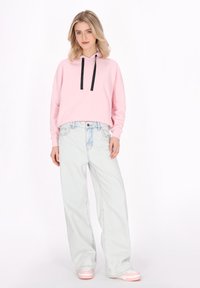 Világos rózsaszínű cropped hoodie fekete húzózsinórokkal, világoskék, bő szárú farmernadrággal és rózsaszín sportcipővel kombinálva, fehér háttér előtt.