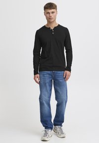 Schwarzes Langarm-Henley-Shirt mit Drei-Knopf-Leiste, kombiniert mit mittelblauen Straight-Leg-Jeans und neutralen Sportschuhen.