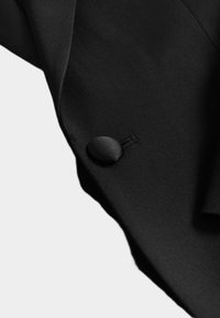 Tela negra con textura suave, que presenta un botón redondo negro y detalles de costura. El diseño muestra un ajuste elegante y entallado.