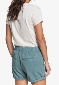 Světle růžové tričko s krátkými rukávy spárované s tyrkysovými šortkami s elastickým pasem a ohrnutými lemy, s dvěma zadními kapsami.