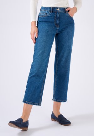 Frau trägt blaue, kurze Jeans, weißes langärmliges Oberteil und marineblaue Loafers, steht vor einem neutralen Hintergrund.