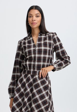 Femme aux longs cheveux foncés portant une robe à manches longues à carreaux marron et blanc, debout avec une main sur la hanche devant un fond uni.