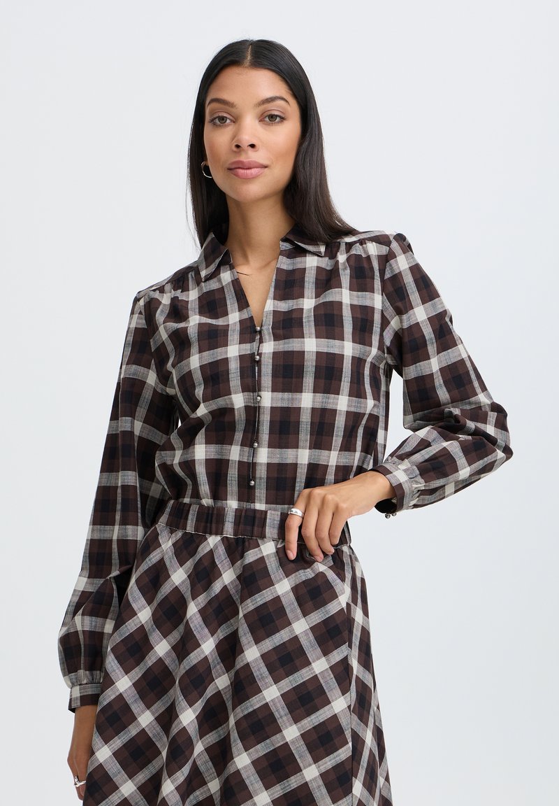 Femme aux longs cheveux foncés portant une robe à manches longues à carreaux marron et blanc, debout avec une main sur la hanche devant un fond uni.