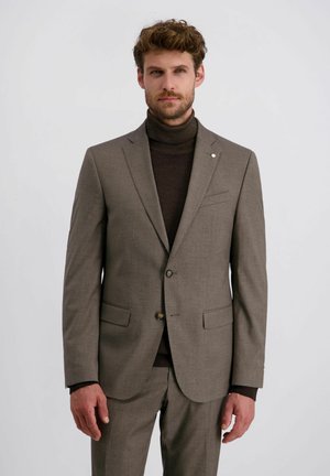 Mann mit Bart, der einen maßgeschneiderten taupefarbenen Anzug und einen dunkelbraunen Rollkragenpullover vor einem schlichten, hellen Hintergrund trägt.