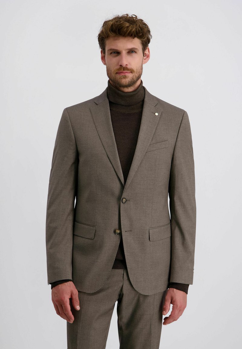 Mann mit Bart, der einen maßgeschneiderten taupefarbenen Anzug und einen dunkelbraunen Rollkragenpullover vor einem schlichten, hellen Hintergrund trägt.