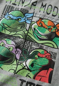 Koszulka z nadrukiem przedstawiająca cztery stylizowane zielone żółwie w różnych kolorach: niebieskim, pomarańczowym, fioletowym i czerwonym, na szarym tle materiału.