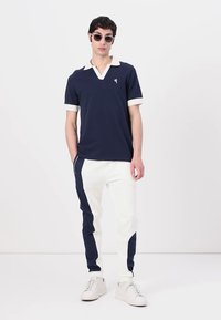 Jūras spēku polo krekls ar baltu apkakli un aprocēm, apvienots ar baltām biksēm ar jūras spēku krāsas sānu svītrām. Modelis valkā saulesbrilles un sporta apavus.