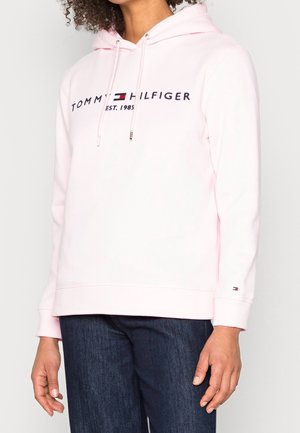 Personne portant un sweat à capuche Tommy Hilfiger rose clair avec logo et texte, associé à un jean bleu foncé, debout devant un fond blanc.