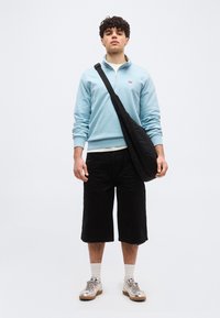 Νεαρός άντρας στέκεται φορώντας ανοιχτό γαλάζιο φούτερ με φερμουάρ μέχρι τη μέση, μαύρα σορτς μέχρι το γόνατο, ασημένια αθλητικά παπούτσια, λευκές κάλτσες και μαύρη τσάντα χιαστί.