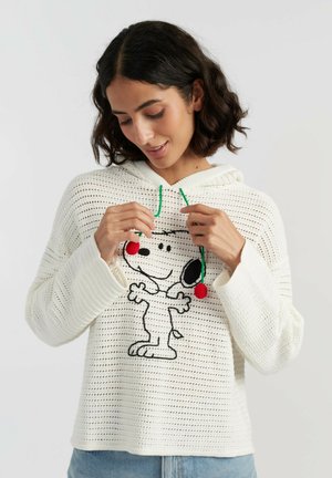 Femme aux cheveux foncés portant un hoodie blanc en maille avec motif Snoopy, tenant des cordons verts avec pompons rouges, sur fond uni.