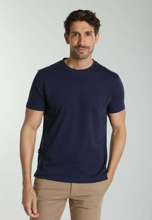 Homme portant un t-shirt bleu marine à manches courtes et un pantalon beige, debout avec une main dans la poche, sur un fond clair.