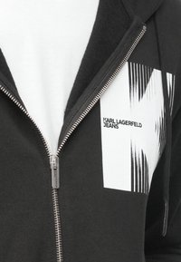 Μαύρο φούτερ με φερμουάρ και λευκό γραφικό που απεικονίζει κάθετες γραμμές και το κείμενο "KARL LAGERFELD JEANS" στην αριστερή πλευρά του στήθους. Βαμβακερό ύφασμα.