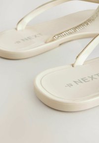 Sandales tongs beige avec des brides fines, une bride décorée de petits strass brillants, sur une surface neutre.