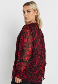 Bluse mit rotem Blumenmuster aus transparenter schwarzer Stoff, langen Ärmeln, gerafften Manschetten und dekorativen Akzenten am Saum. Elegantes und leichtes Design.