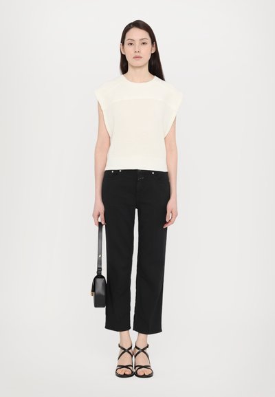Hvit strikkegenser med korte ermer, parret med svarte cropped jeans og svarte sandaler med stropper. Holder en liten svart håndveske.