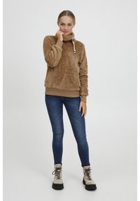Brun fleece pullover med høj krave og snoretræk. Kombiner med blå tætsiddende jeans og brune snørebåndsstøvler med en kraftig sort sål.