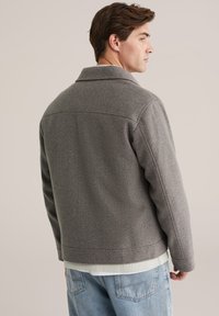 Grijze wollen jas met een kraag, zichtbare stiksels en een rechte snit, gedragen over een wit shirt en lichtblauwe jeans.