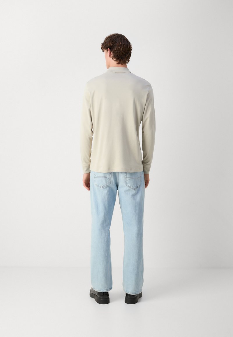 Chemise à manches longues beige clair avec une texture subtile, associée à un jean bleu clair ample et des chaussures noires, vue de derrière.