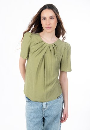 Blouse vert olive avec manches courtes bouffantes et encolure plissée, en tissu léger. Associée à un jean bleu clair.