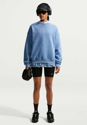 Donna che indossa una felpa blu oversize, pantaloncini da ciclismo neri, cappello nero, occhiali da sole, cuffie, che tiene una borsa nera, in piedi su uno sfondo uniforme.