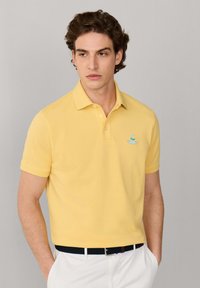 Homme aux cheveux bouclés bruns portant un polo jaune et un pantalon blanc, debout les mains dans les poches devant un fond gris.