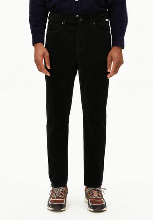 Homme portant un pantalon en velours côtelé noir, une chemise bleu marine et des baskets marron et grises, debout les bras détendus le long du corps.
