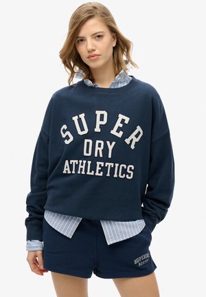 Donkerblauwe sweatshirt met "SUPER DRY ATHLETICS" in crèmekleur, gecombineerd met een blauw gestreept overhemd met kraag en donkerblauwe shorts met logo-detail.
