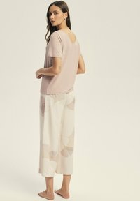 Jasnoróżowy t-shirt z dekoltem w serek, zestawiony z kremowymi spodniami o szerokich nogawkach z jasnym kwiatowym wzorem i miękką teksturą. Bez obuwia.