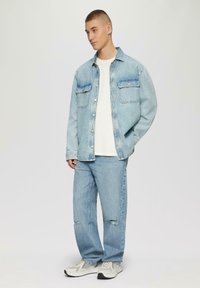 Helle blaue Jeansjacke mit zwei Brusttaschen, kombiniert mit passenden, ausgewaschenen Jeans. Darunter ein weißes T-Shirt und neutrale Sneaker.