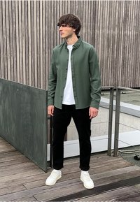 Mladý muž s hnědými vlasy, oblečený v zelené košili na knoflíky přes bílém tričku, černých kalhotách a bílých teniskách, stojí na dřevěné terase.