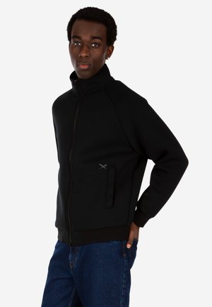 Junge Person trägt eine schwarze Jacke mit Reißverschluss und hohem Kragen sowie blaue Jeans, steht mit einer Hand in der Tasche und blickt nach vorne.