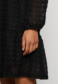 Črna obleka z teksturiranim čipkastim vzorcem, ki vsebuje cvetlične motive, napihnjene rokave in večplastno robno obrobo; posnetek v bližini roke.