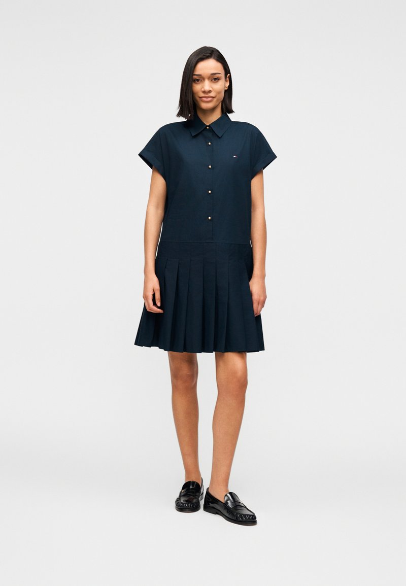 Φόρεμα πουκάμισο σε ναυτικό μπλε χρώμα με κουμπιά μπροστά, κοντά μανίκια και πλισέ φούστα. Φορεμένο με μαύρα loafers, με διακριτική λεπτομέρεια λογότυπου.