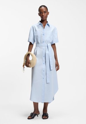 Donna che indossa un abito midi azzurro con bottoni, maniche corte e cintura, che tiene una borsa rotonda intrecciata e porta sandali neri con tacco.