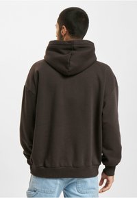 Brun hoodie med snøre i hætten, løs pasform, lange ærmer og ribbede manchetter. Stoffet virker blødt med en glat tekstur.