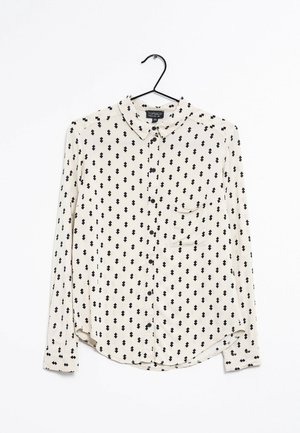Chemise blanche à manches longues boutonnée avec motifs losanges noirs, col et une poche poitrine, suspendue sur un cintre noir sur fond blanc.