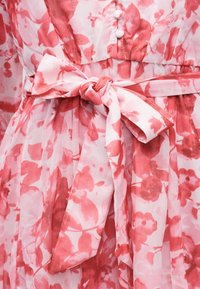 Sarkanrozā ziedu raksta kleitas detaļa ar auduma jostu, kas sasieta lokā pie vidukļa, un baltām pogām uz krūšu daļas.