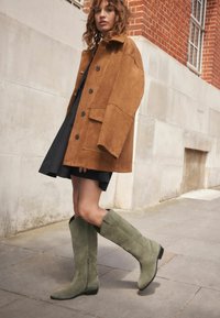 Abrigo oversized de ante marrón con botones grandes, llevado sobre un vestido negro, combinado con botas de ante verde claro hasta la rodilla en una acera.